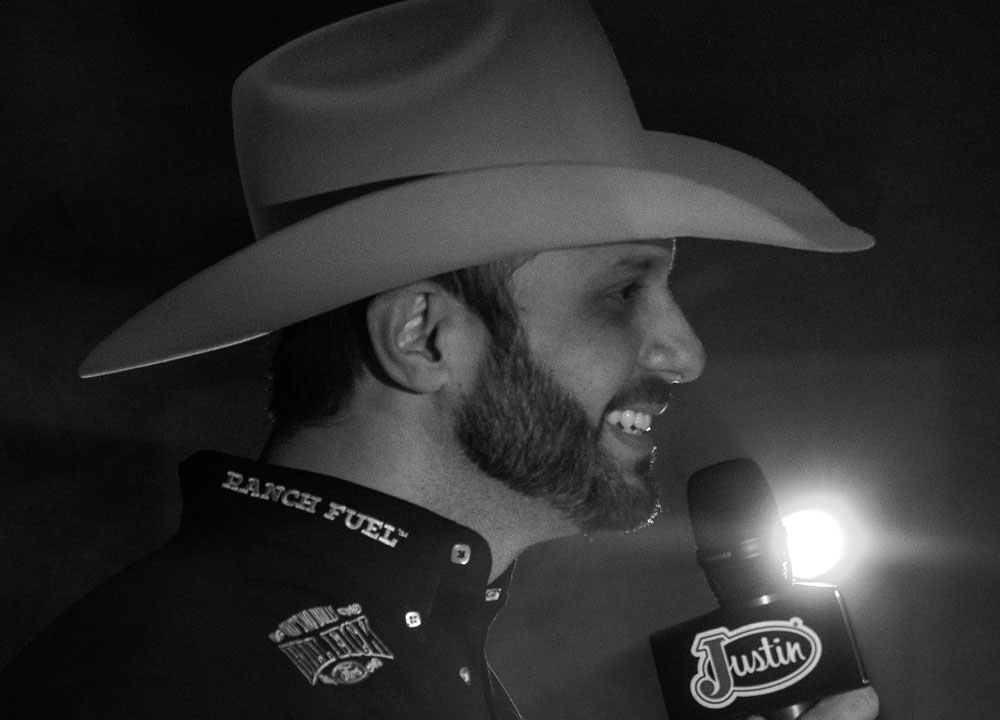 Anthony Lucia vistiendo una camisa negra y un sombrero de vaquero, sosteniendo un micrófono y hablando con una luz detrás de él.